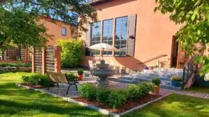 Auringon valaisemassa puutarhassa suihkulähde ja puinen penkki. Taustalla on kermanvärinen rakennus, jossa on suuret ikkunat ja sateenvarjolla peitetty oleskelutila. Rehevä vehreys ja hoidetut pensaat ympäröivät kivipolkua.