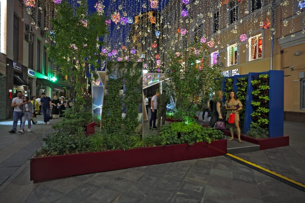 Ryhmä ihmisiä kävelemässä puun ympärillä valojen kanssa. Kuvassa on harmaat värit. Näyttää yhden henkilön. Mukana myös joulukuusi, joulu, rakennus.