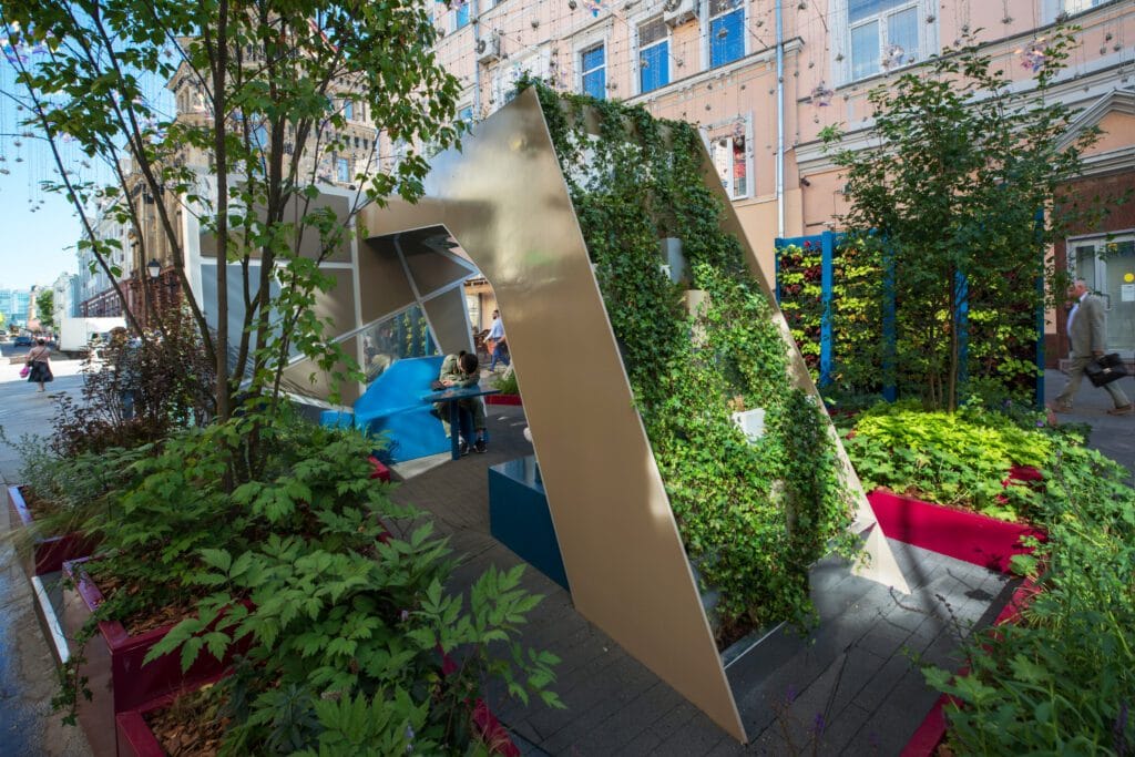 Puutarha, jossa on penkki ja rakennus taustalla. Kuvassa on vihreä ja harmaa väri. Mukana myös ulkona, kasvi, puu.