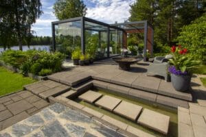 Lasirakennus, jossa on lasinen etuosa. Kuvassa on harmaa ja musta väri. Sisältää kasvin, ruukkukasvin ja penkin. Mukana myös ulkona, taivas, puu.