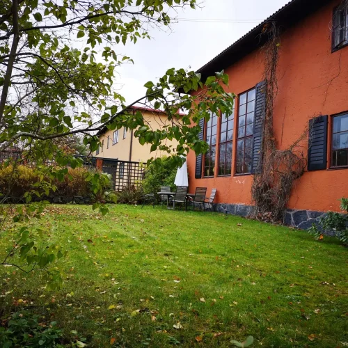 Viihtyisä takapiha, jossa on vihreää ruohoa, puiden ympäröimänä. Oikealla on oranssi talo, jossa on suuret ikkunat ja tummat ikkunaluukut. Kaksi tuolia ja valkoinen sateenvarjo on sijoitettu talon lähelle, mikä viittaa rauhalliseen ulkoistuinalueeseen.