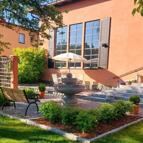 Auringon valaisemassa puutarhassa suihkulähde ja puinen penkki. Taustalla on kermanvärinen rakennus, jossa on suuret ikkunat ja sateenvarjolla peitetty oleskelutila. Rehevä vehreys ja hoidetut pensaat ympäröivät kivipolkua.