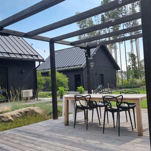 Kansi, jossa on pöytä ja tuolit ja talo taustalla. Kuvassa on harmaa ja musta väri. Mukana myös ulkona, taivas, kasvi.