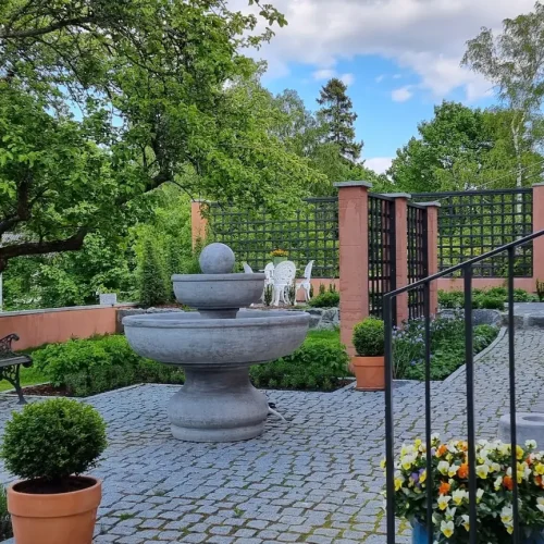 Rauhallinen puutarha, jonka keskellä on kivisuihkulähde, jota ympäröivät ruukkukasvit ja mukulakivipolku. Puinen penkki ja terassituolit istuvat lähellä säleikköä ja oransseja seiniä, ja taustalla on vehreä ja pilvinen taivas.