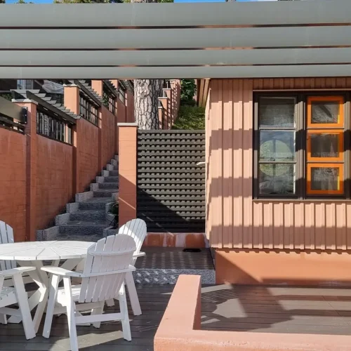 Pieni patio, jossa on pyöreä valkoinen pöytä ja neljä yhteensopivaa tuolia. Tilaa rajaavat lohenväriset seinät, joista ylöspäin johtavat portaat. Yläpuolella on osittain varjossa pergola ja taustalla ikkunallinen rakennus.