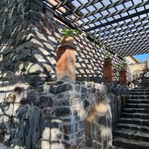 Kiviportaikko ja seinä, jossa puinen pergola heittää ristikkomaisia varjoja. Rakenne on koristeltu kasveilla kulmissa. Jalkakäytävä ja rakennus ovat osittain näkyvissä, taustalla sininen taivas.