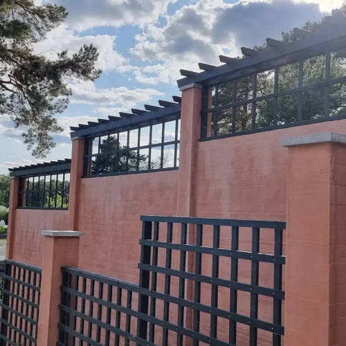 Punatiilinen rakennus, jossa on suuret ikkunat ja ruudukkomaiset metallirakenteet, asetettu taivasta vasten, jossa pilvet ja auringonvalo kurkistaa läpi. Vasemmalla on puu, ja taustalla näkyy joki vehreällä puolella.