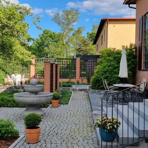 Viehättävä puutarha, jossa on kivipatio, jossa on pyöreä suihkulähde ja ruukkukasveja. Pöytä tuoleineen ja sateenvarjo sijaitsevat taloon johtavien portaiden vieressä. Vihreys ympäröi aluetta kirkkaan sinisen taivaan alla ja pilviä.