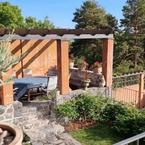 Viehättävä ulkopatio, jossa on pergola, josta on näkymät puille ja veteen. Tilaan kuuluu sininen pöytäliina, kiviseinien ja vehreyden ympäröimä pöytä, jonka etualalla on ruukkukasvi.