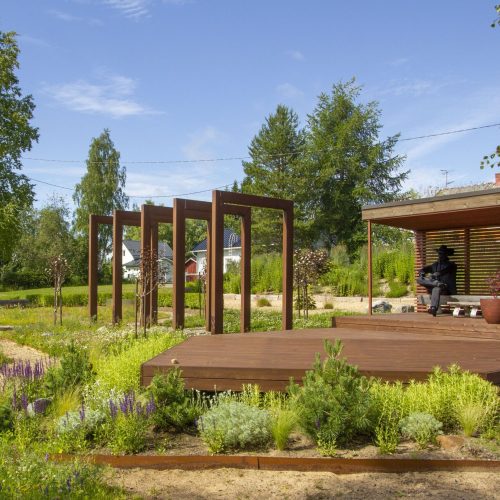 Henkilö, joka istuu penkillä puutarhassa. Kuvassa on ruskeat värit. Sisältää myös ulkona, kasvina, pilvenä.