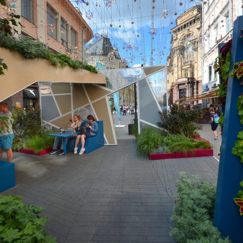 Ryhmä ihmisiä istuu pöydän ääressä kapealla kujalla rakennusten välissä. Kuvassa on harmaat värit. Mukana myös ulkona, kasvi, taivas.