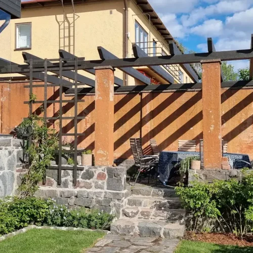 Aurinkoisella takapihalla on kivipatio, jossa puinen pergola luo varjoja maahan. Siellä on ulkoistuinalue raidallisilla tyynyillä ja ruukkukasveilla. Taustalla sinisen taivaan alla on kaksikerroksinen talo kerman ja ruskean seinän kera.