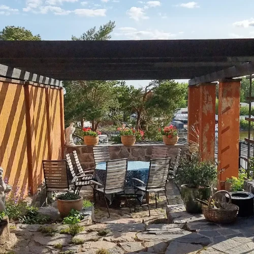 Maalaismaisella ulkopatiolla on puinen pergola, jossa on varjokuvio. Alla on pöytä, jota ympäröivät tuolit. Ruukkukasveja ja kukkia koristavat kivilattiat. Taustalla näkyy puita ja kirkas taivas.