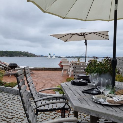 Pöytä ja tuolit vesistön vieressä. Kuvassa on harmaa ja valkoinen väri. Sisältää sateenvarjon, tuolin ja ruokapöydän. Huoneessa on myös ulkona, taivas, huonekalut.