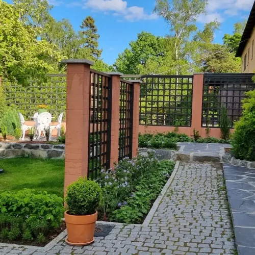 Puutarhamaisema, jossa on mukulakivipolku, ruukkukasveja ja patio, jossa on tuolit ja sateenvarjo. Aluetta ympäröi vehreys ja säleiköt, taustalla kivisuihkulähde ja kaksikerroksinen talo kirkkaan sinisen taivaan alla.