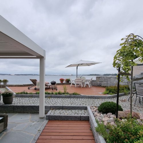 Patio, jossa on pöytä ja tuolit ja sateenvarjot vesistön äärellä. Kuvassa on harmaa ja valkoinen väri. Sisältää tuolin, sateenvarjon ja kasvin. Mukana myös ulkona, taivas, pilvi.