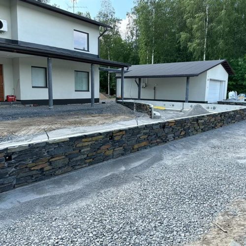 Kivimuuri rakennuksen vieressä. Kuvassa on harmaat värit. Mukana myös ulkona, maa, puu.