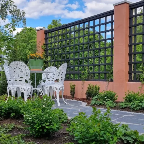 Patio, jossa on valkoinen koristeellinen metallipöytä ja kaksi tuolia, jotka on asetettu kivilaatoille. Takana on koristeellinen ristikkoaita, jossa kasvit kiipeilevät. Vihreät pensaat ja pienet puut ympäröivät aluetta, ja sininen taivas pilvineen on näkyvissä.