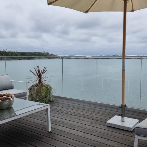 Kansi, jossa on pöytä ja tuolit sekä vesistö taustalla. Kuvassa on harmaa ja valkoinen väri. Sisältää vuodesohvan ja ruukkukasvin. Huoneessa on myös taivas, ulkona, sohvapöytä.