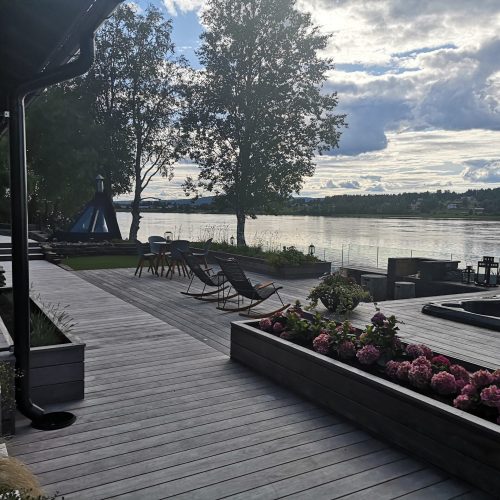 Kansi, jossa on tuoleja, pöytiä ja kukkia vesistön vieressä. Kuvassa on harmaa ja musta väri. Mukana myös pilvi, ulkona, taivas.