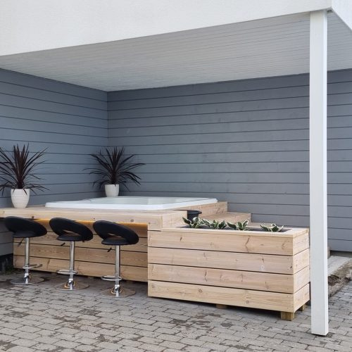 Koira istuu talon ulkopuolella. Kuvassa on harmaa ja valkoinen väri. Sisältää ruukkukasveja, -kasveja ja -nisäkkäitä. Huoneessa on myös ulkona, rakennuksena, huonekaluina.