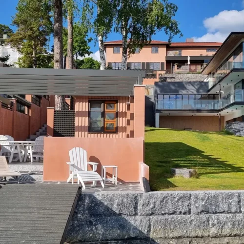 Moderni huvila, jossa on suuret ikkunat rinteessä, ja siinä on monitasoinen suunnittelu. Etualalla on pergolalla peitetty patio tuoleineen ja pöytineen, nurmialue ja kivipolku. Puut ja kirkas sininen taivas taustalla.