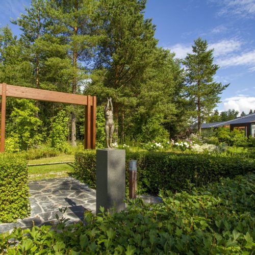 Puutarha, jonka taustalla on talo. Kuvassa on vihreitä värejä. Näyttää yhden henkilön. Mukana myös ulkona, pilvi, taivas.