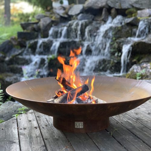 Tulikuoppa liekillä. Kuvassa on harmaa ja musta väri. Huoneessa on myös ulkona, kasvi, takka.