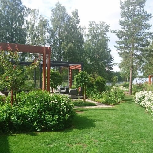 Puutarha, jossa on huvimaja. Kuvassa on vihreä ja harmaa ja valkoinen väri. Sisältää puuta ja kasvia. Huoneessa on myös ulkona, ruohoa, maisemointia.