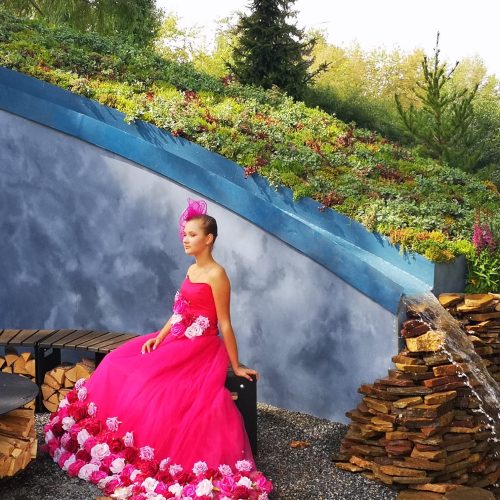 Vaaleanpunaisessa mekossa oleva henkilö seisoo vesiputouksen edessä. Näyttää yhden henkilön. Mukana myös vaatteet, ulkona, puu.