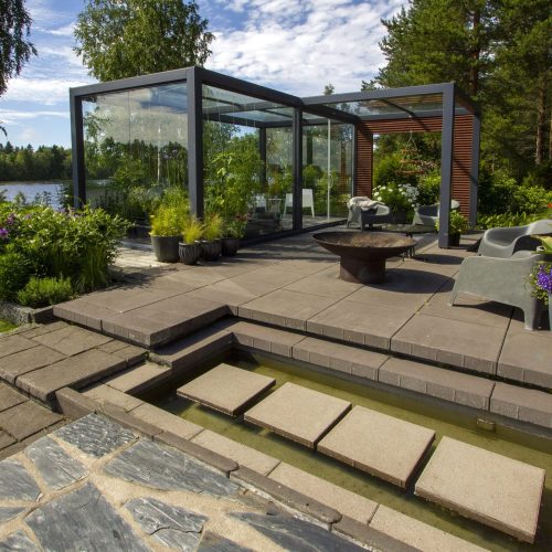 Lasirakennus, jossa on lasinen etuosa. Kuvassa on harmaa ja musta väri. Sisältää kasvin, ruukkukasvin ja penkin. Mukana myös ulkona, taivas, puu.